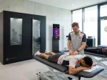 Fußballspieler wird auf einer Liege von einem Physiotherapeuten behandelt. Im Hintergrund ist die Kältekammer zu sehen